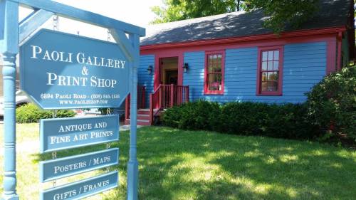 A blue building with red trim houses the Paoli Gallery & Print Shop, featuring a sign with its name and services.