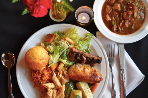 A plate of food featuring rice, salad, cornbread, pasta, and a side of stew, with flowers and a candle in the background.