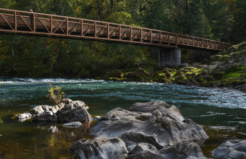 A wooden bridge spans a river, surrounded by lush greenery and rocky banks under a clear blue sky.