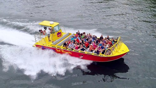 A yellow and red jet boat speeds across the water, filled with excited passengers enjoying the ride.