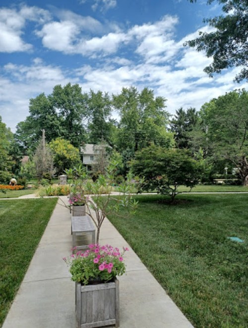 A pathway lined with flower planters, leading through a lush green garden under a partly cloudy sky.