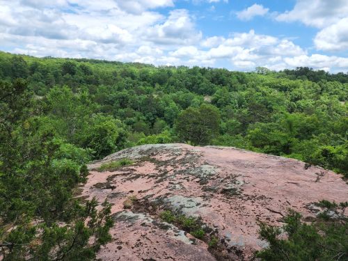 A rocky outcrop overlooks a lush green forest under a partly cloudy sky.