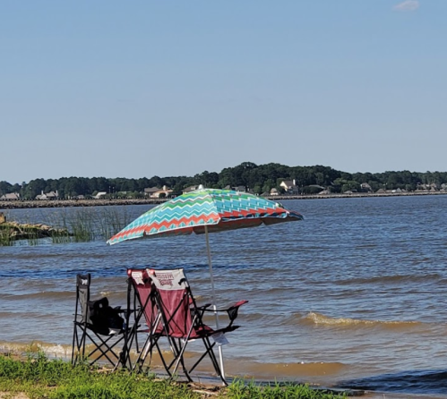 Two beach chairs under a colorful umbrella by a calm lake, with trees and houses in the background.