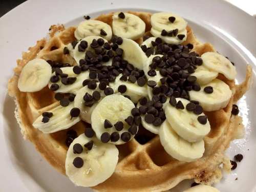 A golden waffle topped with sliced bananas and chocolate chips on a white plate.