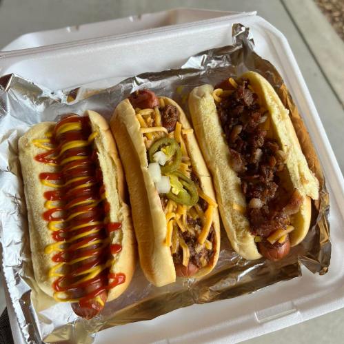 Three hot dogs in a takeout container: one with ketchup, one with jalapeños and cheese, and one with bacon bits.