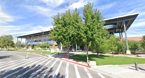 Modern building with large windows, surrounded by trees and a landscaped area, under a blue sky with clouds.