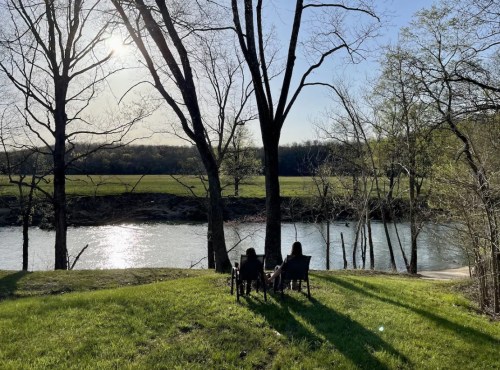 Two people sit in chairs by a river, surrounded by trees, enjoying a sunny day with a scenic view.