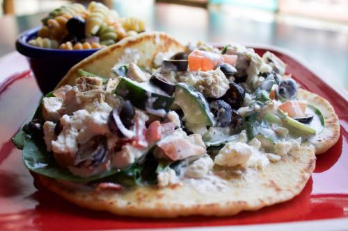 A colorful plate featuring a salad on flatbread with vegetables, chicken, olives, and a side of pasta salad.