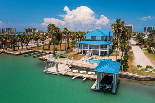 Aerial view of a blue-roofed house by the water, featuring a pool, dock, and palm trees under a clear blue sky.