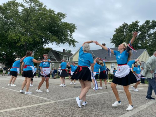 A group of women in blue shirts and skirts dance joyfully in a street, celebrating a festive event.