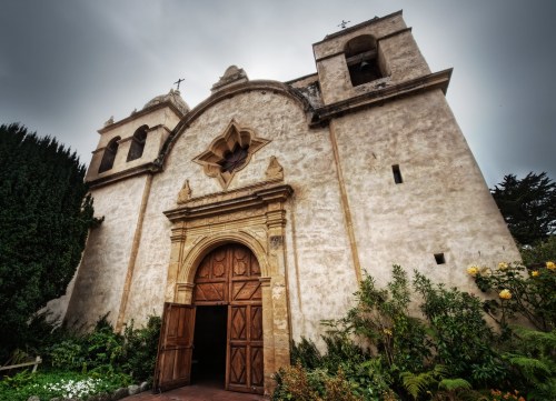 Historic church with a large wooden door, ornate window, and surrounding greenery under a cloudy sky.