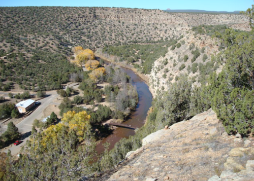 A winding river flows through a rocky landscape, surrounded by trees and hills under a clear blue sky.