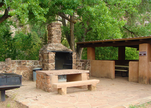 A stone barbecue grill and picnic area surrounded by trees in a park setting.