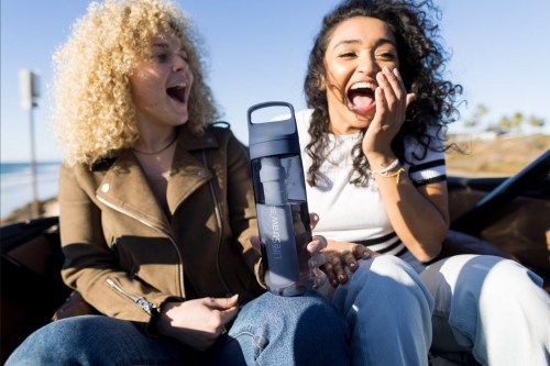 Two women laughing together outdoors, holding a water bottle, with a beach in the background.