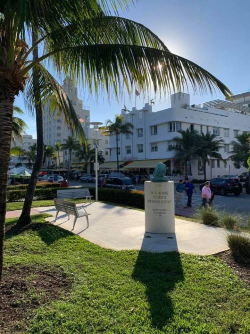 A sunny street scene in South Beach, Miami, featuring palm trees, a bench, and a monument with a shadow.