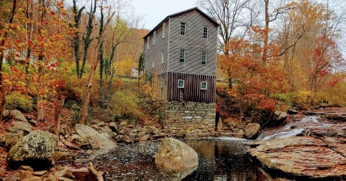 A historic wooden mill stands by a stream, surrounded by colorful autumn foliage and rocky terrain.