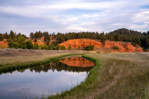 A serene landscape featuring a winding river reflecting red rock formations and green trees under a cloudy sky.