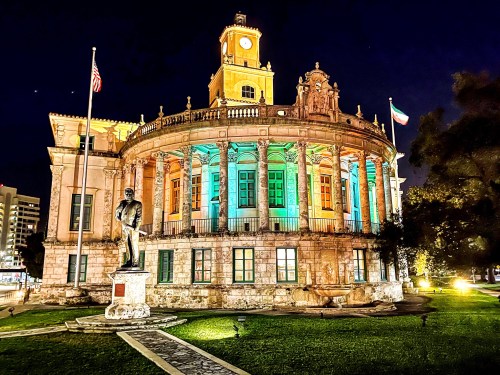 Historic building illuminated at night, featuring a clock tower and a statue in front, surrounded by greenery.