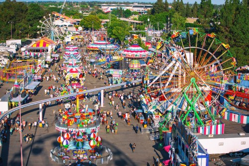 A vibrant fairground scene with colorful rides, games, and crowds enjoying a sunny day at a carnival.