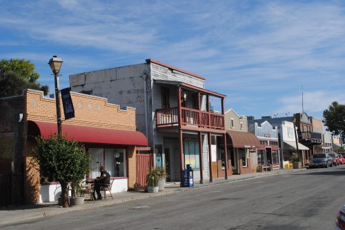 A quiet street scene featuring historic buildings, a lamppost, and a person sitting outside a café under a blue sky.