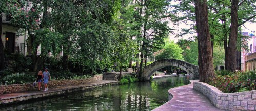 A serene canal scene with lush greenery, a stone bridge, and people walking along the path beside the water.