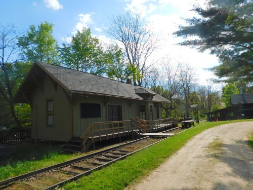 A quaint, wooden building with a ramp, surrounded by trees, alongside a dirt path and old railway tracks.