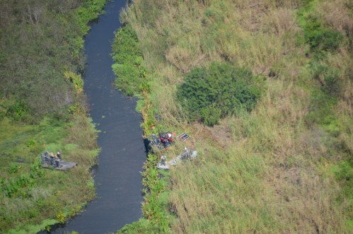 Aerial view of a narrow river surrounded by tall grass, with people working on the bank and a small boat nearby.