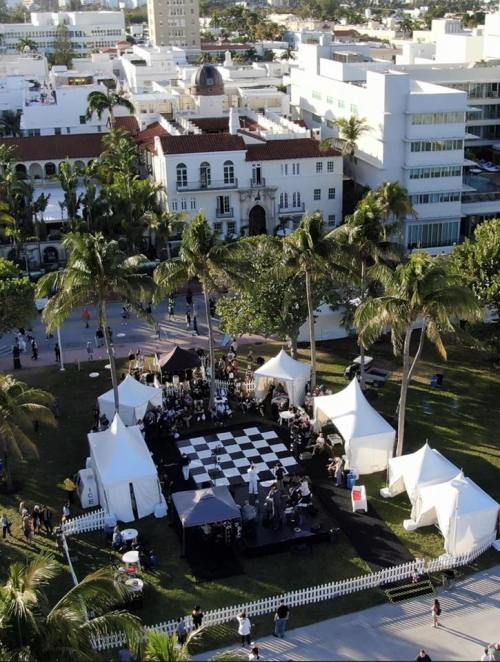 Aerial view of a large outdoor event with a checkered stage, tents, and palm trees in a vibrant urban setting.