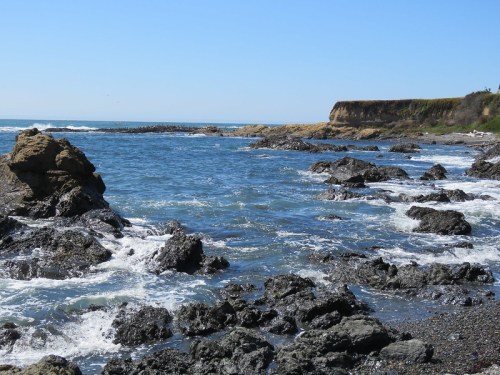 A rocky shoreline with waves crashing against the rocks, under a clear blue sky and distant cliffs.