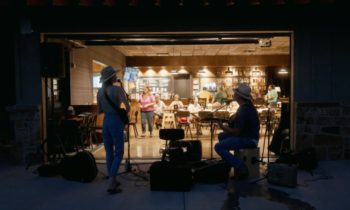 A musician performs outdoors while diners enjoy their meals inside a restaurant, creating a lively atmosphere.