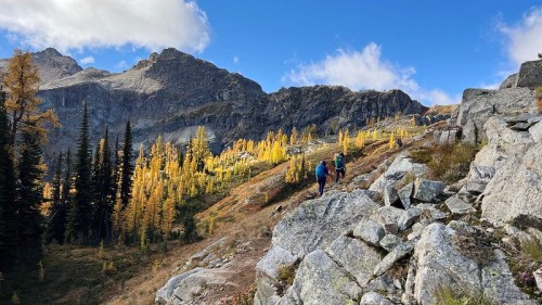 Two hikers navigate a rocky trail surrounded by vibrant autumn foliage and rugged mountains under a blue sky.