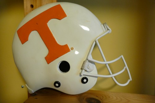 A white football helmet with an orange "T" logo, displayed on a wooden shelf against a yellow background.