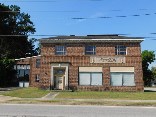 Historic brick building with Coca-Cola signage, featuring large windows and a front entrance, surrounded by greenery.