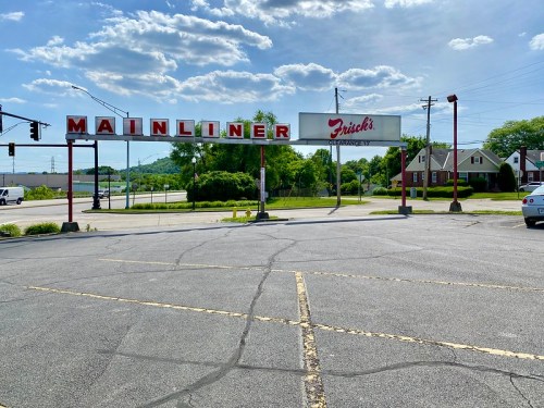 Sign for "Mainliner" with "Frisch's" below, set against a clear blue sky and green landscape. Parking lot in foreground.