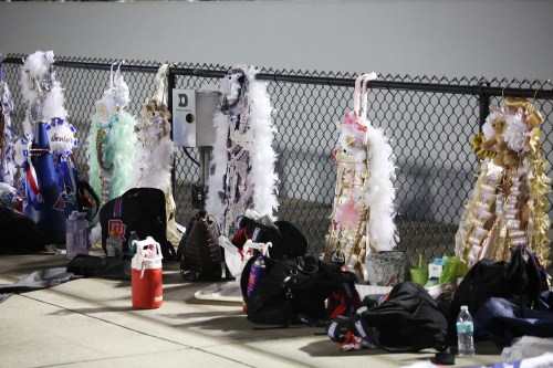 Colorful costumes and bags lined up against a chain-link fence at an event, with a few water bottles visible.