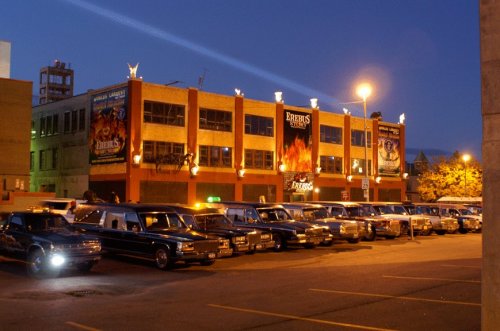 A parking lot at dusk filled with various limousines in front of a brightly lit building with promotional banners.