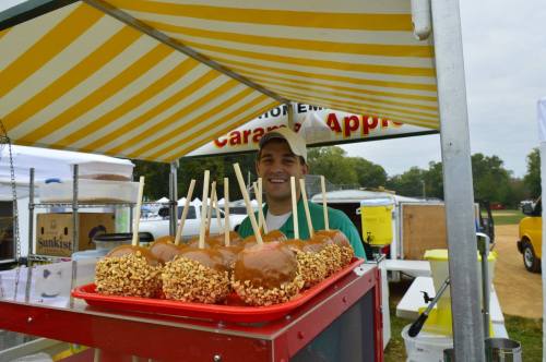 A vendor smiles behind a cart displaying caramel apples topped with nuts at an outdoor event.