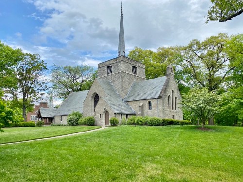 A stone church with a tall spire, surrounded by green trees and grass under a partly cloudy sky.