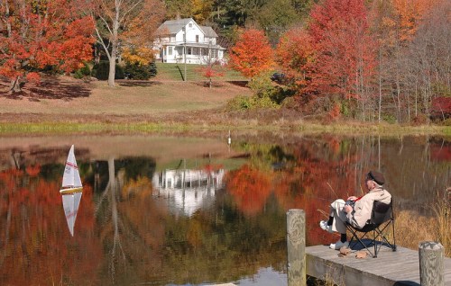 A person sits by a lake, watching a toy sailboat, surrounded by vibrant autumn foliage and a white house in the background.
