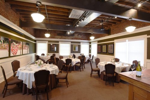 Elegant dining room with round tables, white tablecloths, and artwork on the walls, featuring warm lighting and wooden beams.