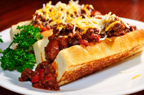 Two chili cheese hot dogs topped with shredded cheese, served on toasted buns with a side of parsley.