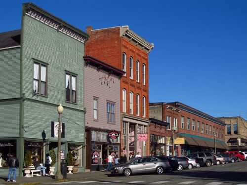 Historic buildings line a sunny street, featuring shops and pedestrians, with clear blue skies above.