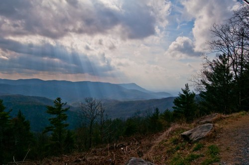 A scenic mountain view with rays of sunlight breaking through clouds, surrounded by trees and rolling hills.