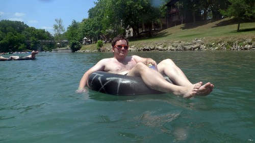 A man relaxes on a black inner tube in a river, wearing sunglasses, with trees and buildings in the background.