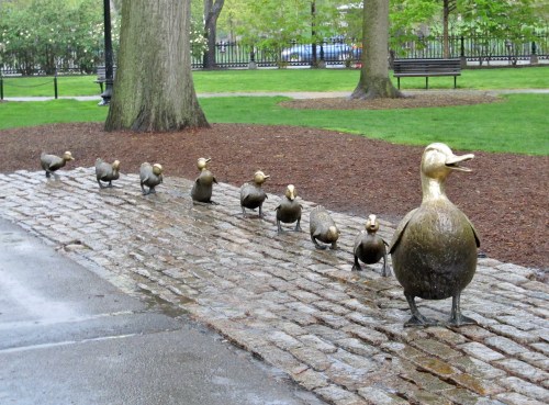 A bronze sculpture of a mother duck and her ducklings walking along a cobblestone path in a park.