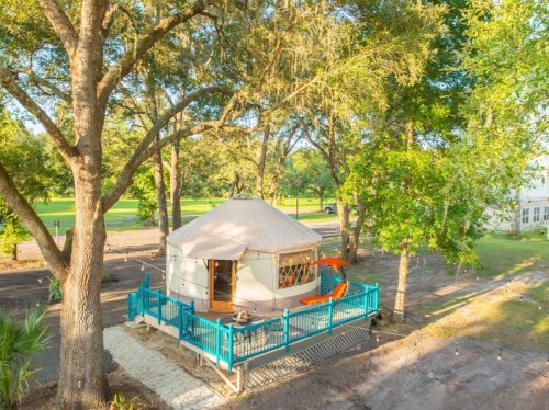 A cozy yurt surrounded by trees, featuring a deck with teal railings and a pathway leading to it.