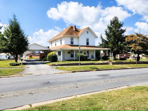 A large white house with a brown roof, surrounded by greenery, on a sunny day with blue skies and fluffy clouds.