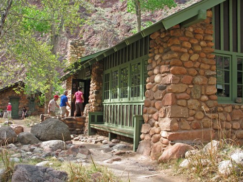 A rustic stone building surrounded by greenery, with people entering and walking nearby in a natural setting.