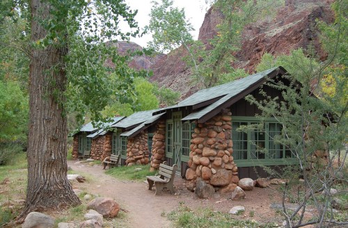 Rustic stone cabins nestled among trees, with a dirt path leading through a scenic landscape of red rock formations.