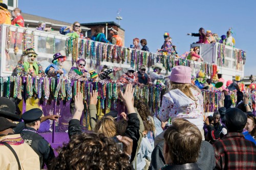 A lively parade scene with colorful floats, people in costumes, and crowds celebrating with beads and festive attire.